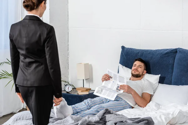 Freundin im Anzug und Freund im Pyjama mit Zeitung, die sich am Wochentag morgens im Schlafzimmer anschaut, Gleichstellungskonzept — Stockfoto