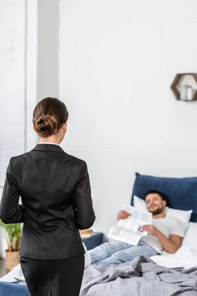 Girlfriend standing in suit and boyfriend in pajamas reading newspaper in bedroom in morning, gender equality concept — Stock Photo