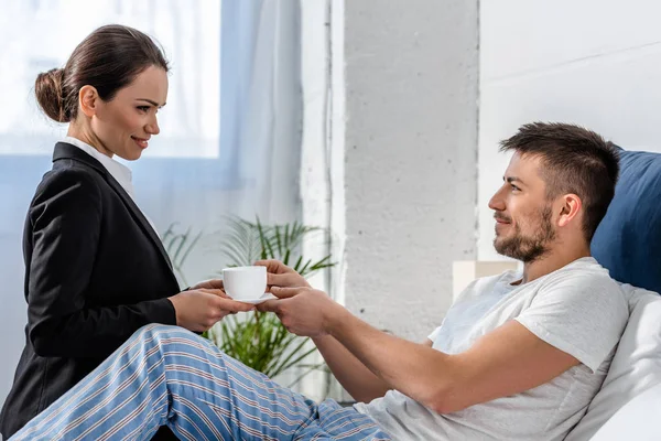 Vista lateral da namorada no terno dando xícara de café ao namorado em pijama no quarto de manhã, conceito de papel social — Fotografia de Stock
