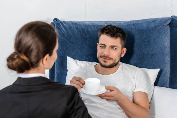 Freundin im Anzug schaut Freund im Pyjama an, morgens Tasse Kaffee im Schlafzimmer, soziales Rollenkonzept — Stockfoto