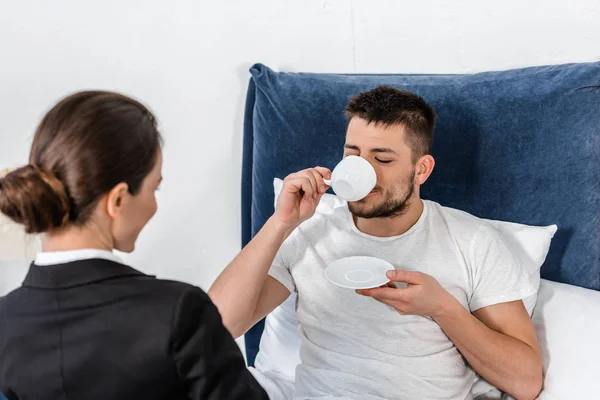 Girlfriend in suit sitting on bed near boyfriend in pajamas drinking coffee in morning at weekday, sexism concept — Stock Photo