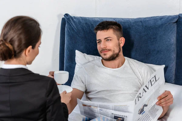 Girlfriend in suit giving cup of coffee to handsome boyfriend in pajamas in bedroom in morning, social role concept — Stock Photo