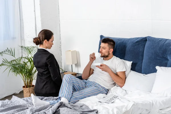 Namorada de terno sentado na cama perto namorado de pijama beber café de manhã no dia da semana, conceito de estereótipos de gênero — Fotografia de Stock