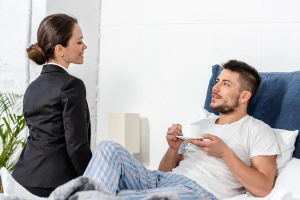 Smiling girlfriend in suit sitting on bed near boyfriend in pajamas drinking coffee in morning at weekday, social role concept — Stock Photo