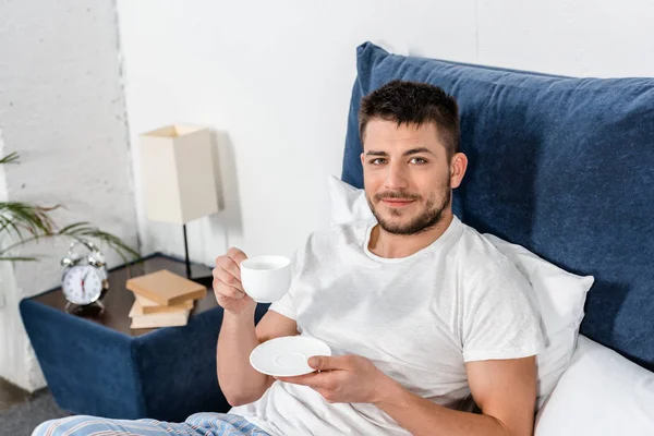 Vista de ángulo alto del hombre guapo sosteniendo la taza de café en la mañana en el dormitorio y mirando a la cámara - foto de stock