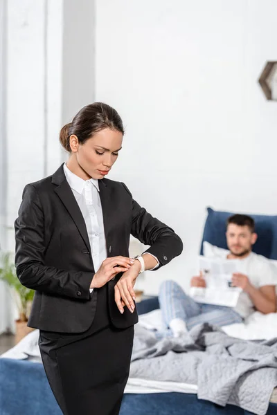 Jolie petite amie en costume vérifier l'heure à la montre-bracelet et petit ami en pyjama lecture journal dans la chambre le matin, concept d'égalité des sexes — Photo de stock