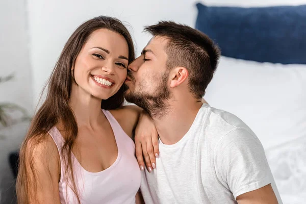 Парень в пижаме обнимает и целует улыбающуюся девушку на кровати в спальне — стоковое фото