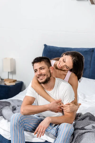 Подружка обнимает улыбающегося парня в пижаме на кровати в спальне — стоковое фото