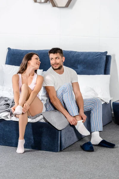 Улыбающаяся девушка и парень в пижаме сидят на кровати и носят носки в спальне — стоковое фото