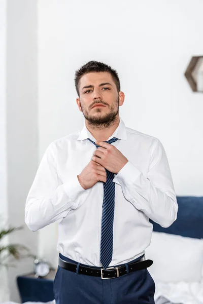 Homem bonito amarrando gravata de manhã no quarto e olhando para a câmera — Fotografia de Stock