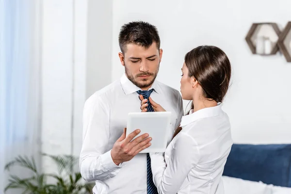 Namorada amarrando gravata namorado enquanto ele usa tablet de manhã em casa, conceito de papel social — Fotografia de Stock