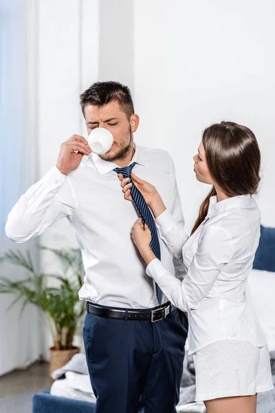 Petite amie attacher petit ami cravate pendant qu'il boit du café le matin à la maison, concept de rôle social — Photo de stock