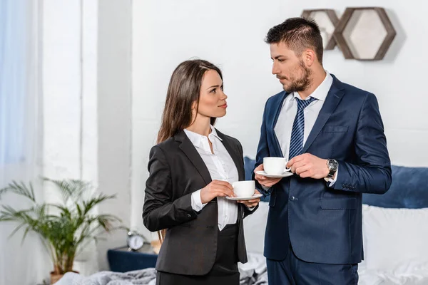Girlfriend and boyfriend in suits holding cups of coffee in morning at home and looking at each other — Stock Photo