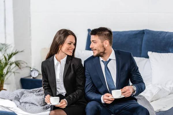 Smiling girlfriend and boyfriend in suits sitting on bed and holding cups of coffee in morning at home, gender equality concept — Stock Photo