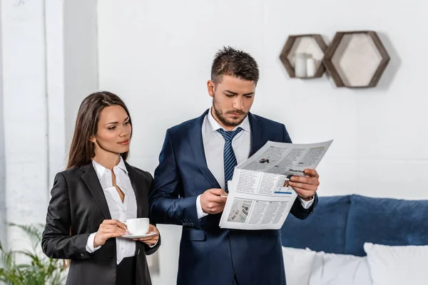 Girlfriend and boyfriend in suits holding cups of coffee in morning, man reading newspaper at home, gender equality concept — Stock Photo