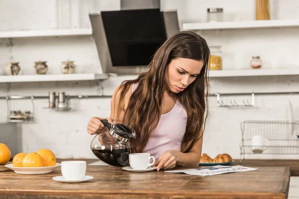 Mulher atraente lendo jornal de negócios e derramando café em xícara de manhã na cozinha — Fotografia de Stock