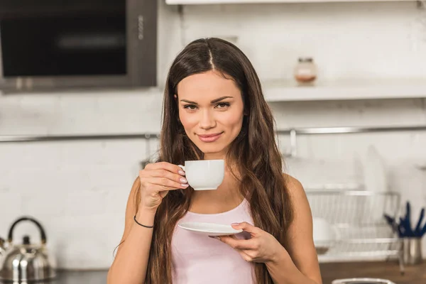 Hermosa mujer alegre sosteniendo la taza de café y mirando a la cámara por la mañana en la cocina - foto de stock