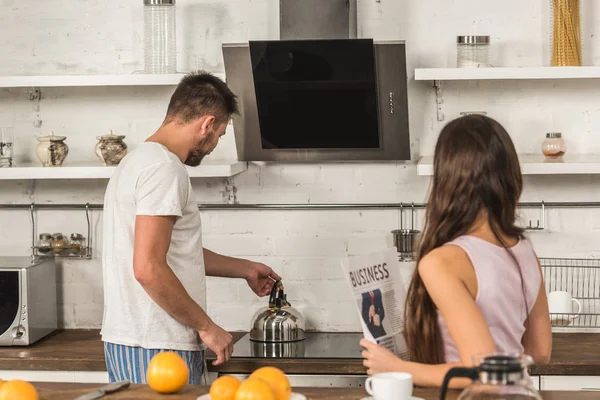 Подружка держит деловую газету и парень ставит чайник на плиту дома — стоковое фото