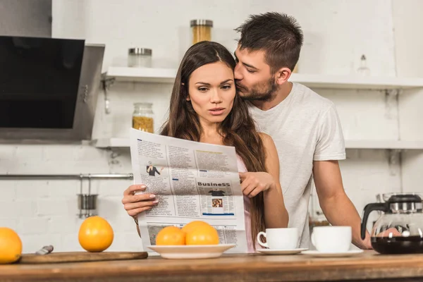 Шокированная девушка читает газету и ее парень целует ее утром на кухне — стоковое фото