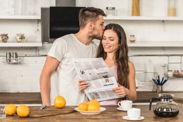 Sonriente novia leyendo periódico y novio besándola en la mañana en casa - foto de stock