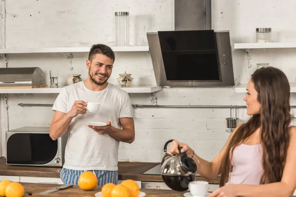 Smiling girlfriend and boyfriend drinking coffee and looking at each other in morning at kitchen — Stock Photo