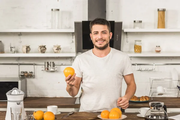 Bell'uomo sorridente che tiene arancione e guarda la macchina fotografica al mattino in cucina — Foto stock