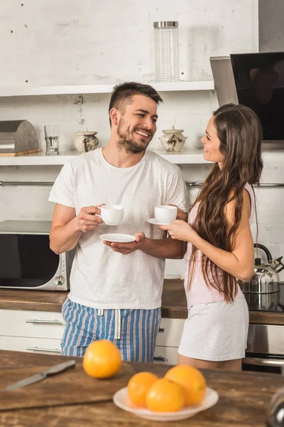 Fidanzata sorridente e fidanzato che tiene in mano tazze di caffè e si guarda al mattino in cucina — Foto stock