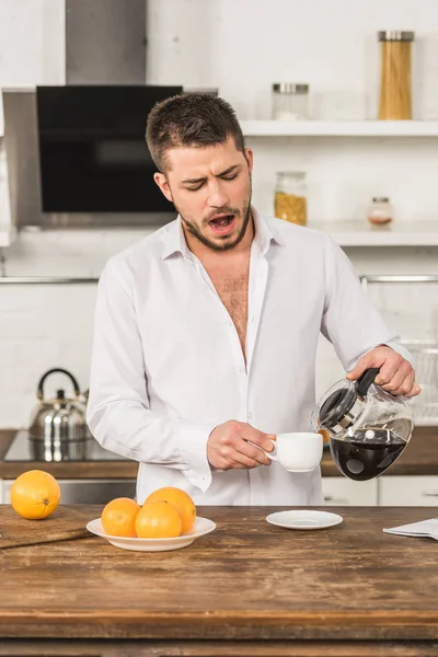 Bell'uomo che sbadiglia e versa caffè in tazza al mattino in cucina — Foto stock