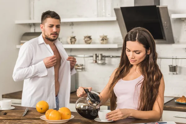 Petit ami boutonnage chemise et petite amie verser du café dans la tasse à la maison, concept de rôles sociaux — Photo de stock