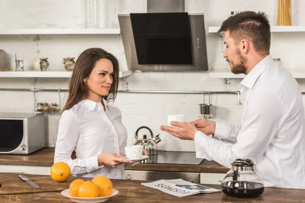 Cheerful boyfriend and girlfriend in shirts standing with cups of coffee in morning at kitchen, gender equality concept — Stock Photo