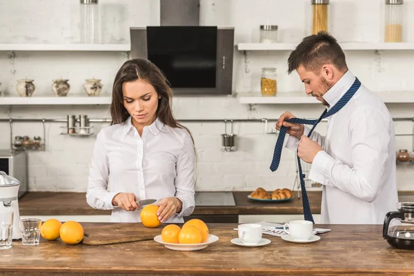 Novio atar corbata y novia corte naranjas en la mañana en la cocina - foto de stock