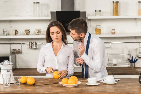 Novio buttoning puño y novia corte naranjas en la cocina, social roles concepto - foto de stock