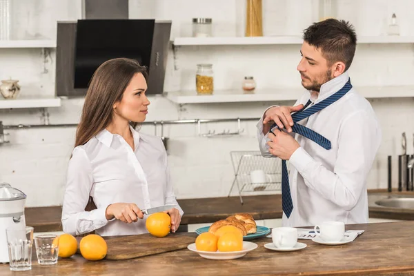 Boyfriend tying tie and girlfriend cutting oranges in morning at kitchen, gender stereotypes concept — Stock Photo
