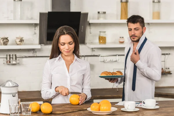 Namorado segurando prato com croissants e namorada cortando laranjas em casa — Fotografia de Stock