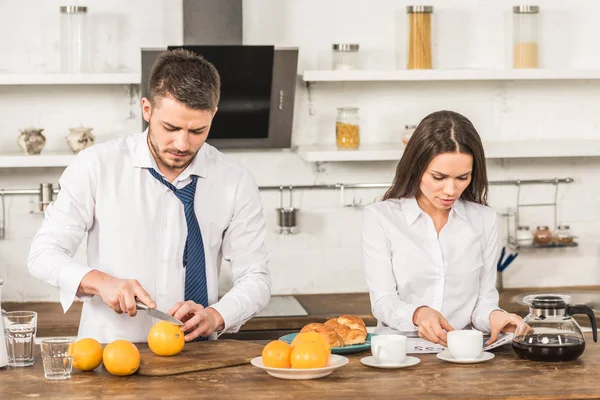 Boyfriend cutting oranges and girlfriend reading newspaper at home, gender equality concept — Stock Photo