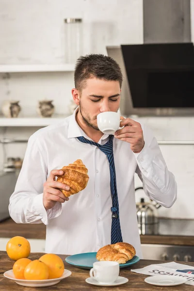 Schöner Mann trinkt Kaffee und isst morgens in der Küche Croissant — Stockfoto