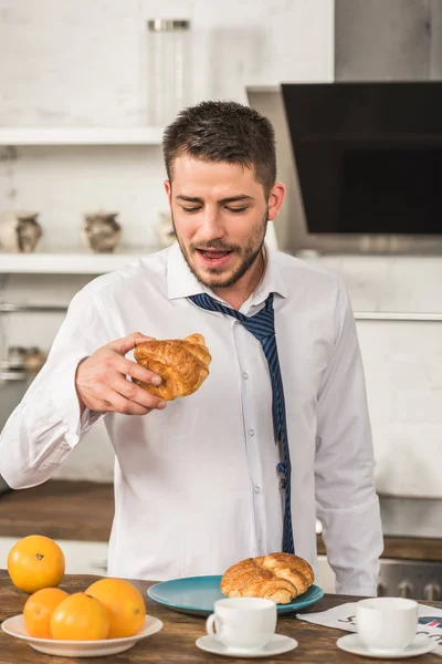 Homem bonito comer croissant de manhã na cozinha — Fotografia de Stock