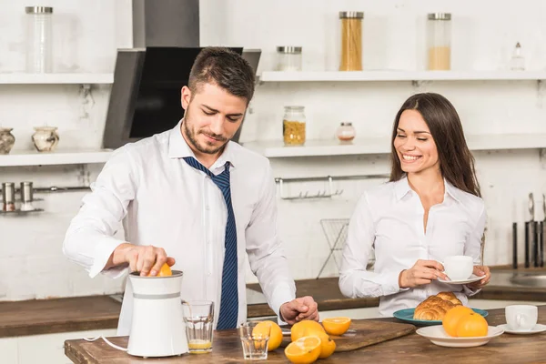 Boyfriend making orange juice and girlfriend standing with cup of coffee in kitchen — Stock Photo