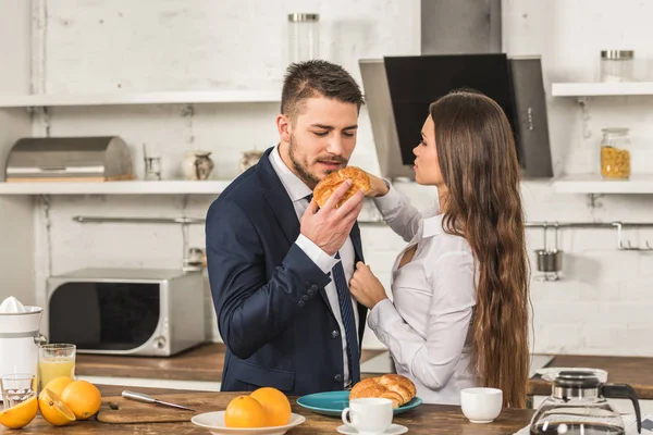 Freund isst Croissant und Freundin repariert seine Jacke in der Küche, Sexismus-Konzept — Stockfoto