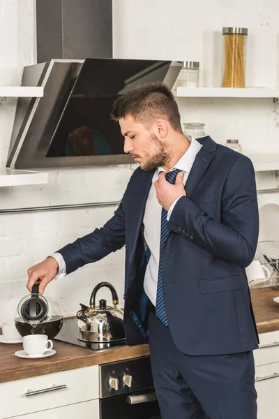 Handsome man in suit pouring coffee into cup and tying tie in morning at kitchen — Stock Photo