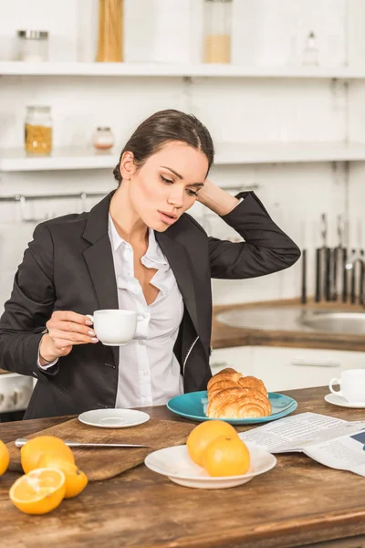 Mulher atraente em terno segurando xícara de café e tocando o cabelo de manhã na cozinha — Fotografia de Stock