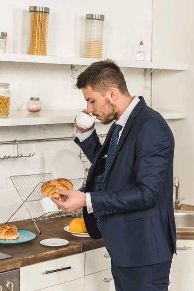 Vista lateral del hombre guapo en traje beber café y la celebración de croissant en la mañana en la cocina - foto de stock
