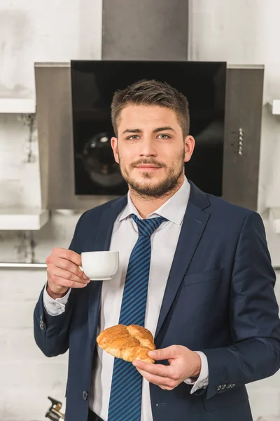 Красивый мужчина в костюме держит чашку кофе и круассан и смотрит в камеру утром на кухне — стоковое фото