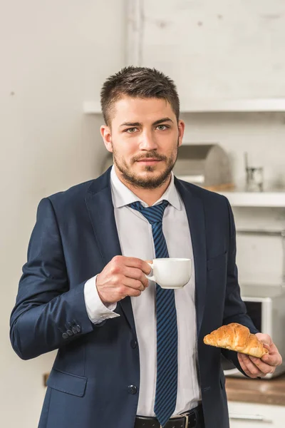 Retrato de hombre guapo en traje sosteniendo taza de café y croissant y mirando a la cámara en la mañana en la cocina - foto de stock