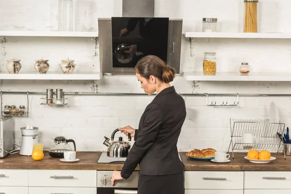Attrayant femme en costume mettant bouilloire sur cuisinière le matin à la cuisine — Photo de stock