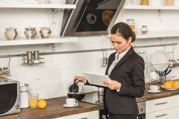 Atractiva mujer vertiendo café en la taza y mirando la tableta por la mañana en la cocina - foto de stock