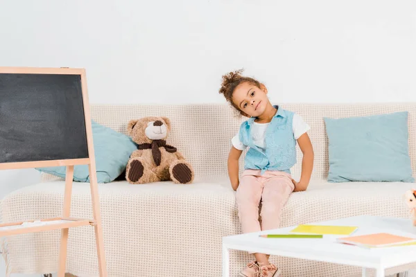 Linda criança afro-americana sentada no sofá e sorrindo para a câmera — Fotografia de Stock