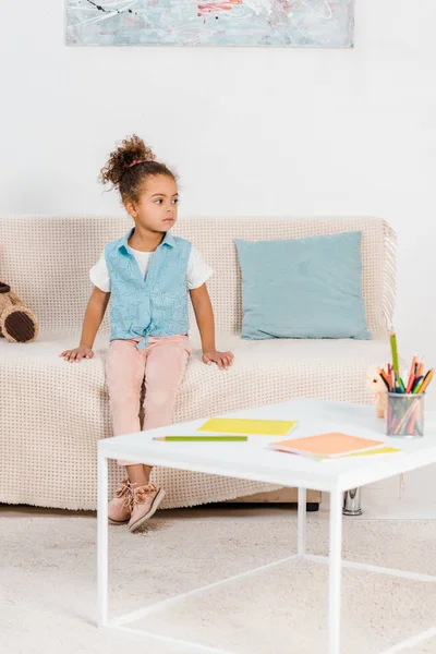 Vue pleine longueur de l'adorable enfant afro-américain assis sur le canapé et regardant loin — Photo de stock