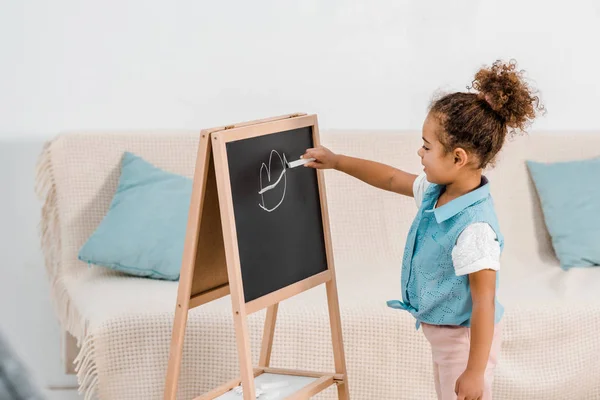 Adorable african american child standing and drawing on chalkboard — Stock Photo