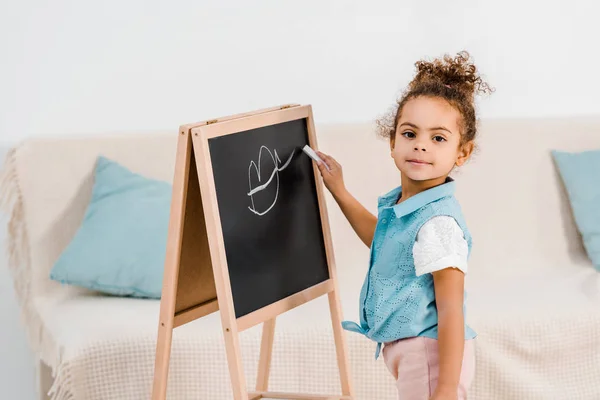 Mignon afro-américain enfant dessin sur tableau noir et regarder la caméra — Photo de stock